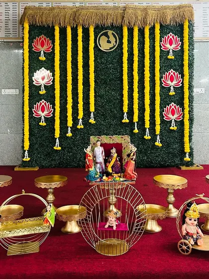 A traditional Seemantham setup featuring a greenery wall with hanging marigold garlands and lotus cutouts. The foreground is arranged with offering tables and decorative cradles.