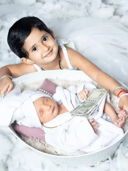 A playful and unique newborn photo with an older sister looking over her baby sibling, who is holding a stack of money.