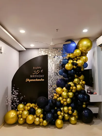 A chic and modern 30th birthday decoration for a home. The setup uses a black and gold color scheme with an organic balloon garland and a custom backdrop with a shimmer panel.