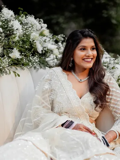 The bride looks radiant and happy, seated on the Sangeet stage surrounded by beautiful white floral arrangements.