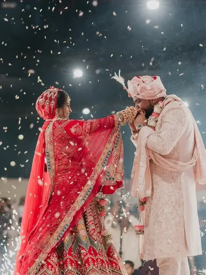 A tender moment during the varmala, where the groom kisses the bride's hand, captured amidst a shower of confetti and sparklers.