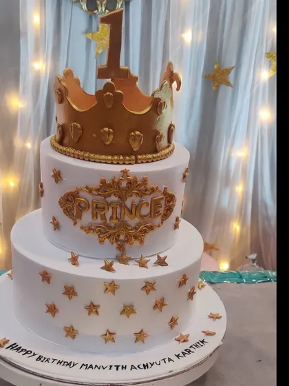 A beautiful three-tiered prince cake in white and gold. The cake is adorned with a large crown, gold stars, and an elegant script, fit for royalty.