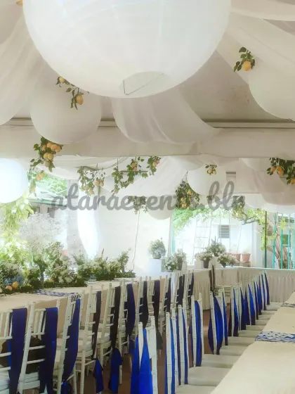 A long view of the Sicilian-themed table setup. The crisp white and blue color scheme, combined with the draped ceiling and hanging lanterns, creates a sophisticated and cohesive look.