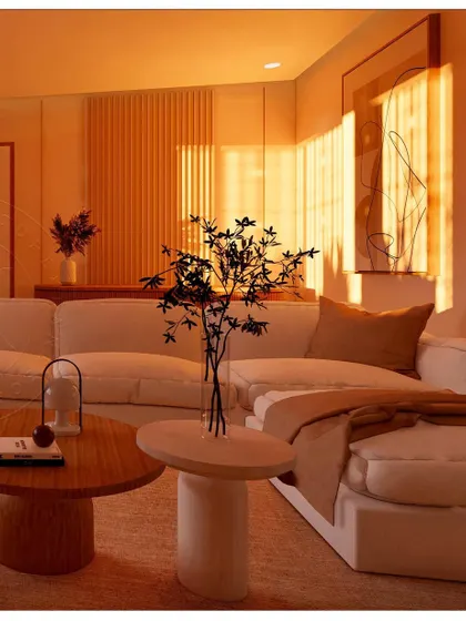 This living room from the "Sharada" project is bathed in the golden glow of the setting sun. The design features a comfortable white sectional sofa, natural wood coffee tables, and subtle textures. The strategic use of lighting, both natural and artificial, creates a warm and inviting ambiance.