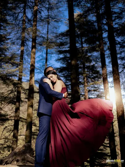 An intimate embrace between a couple in a forest, with the woman's maroon gown creating a striking visual against the sunlit trees.