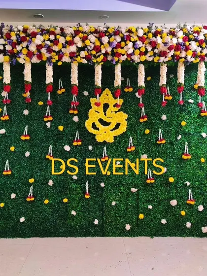 A beautiful backdrop for a housewarming ceremony or engagement. It features a green grass wall, a Ganesha motif made of yellow flowers, and hanging floral strings, creating a divine and welcoming atmosphere.