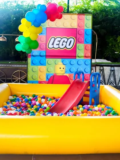 A bright yellow inflatable ball pit complete with a small slide, themed for a LEGO party. It's a safe and fun play area for toddlers and young children.
