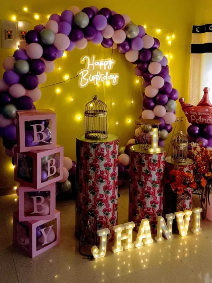 A princess-themed first birthday party for a home celebration. The decor features a purple balloon arch, 'BABY' blocks, birdcage props, and marquee letters.