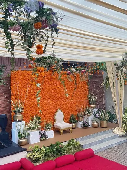A full view of a beautiful outdoor Haldi setup. The vibrant marigold wall provides a pop of traditional color, while the hanging floral chandeliers and draped ceiling add a touch of modern elegance.