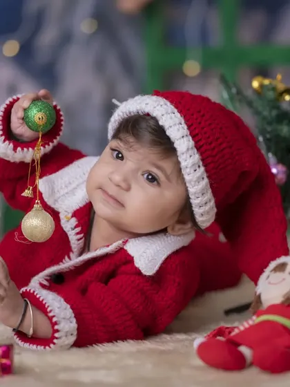 An adorable baby in a hand-knitted Santa outfit, playing with Christmas baubles. The soft, cozy setting is perfect for capturing tender holiday moments.