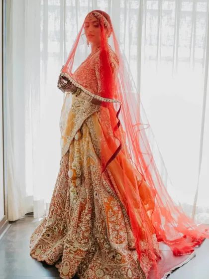 A full-length bridal portrait showcasing the bride's stunning orange and red lehenga. The light from the window beautifully illuminates her, creating a classic and elegant shot.