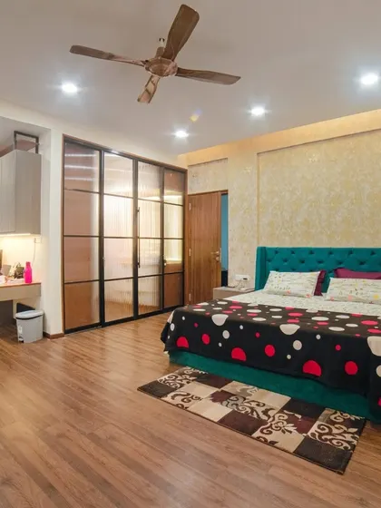 The master bedroom from Ms. Megha Joshi's home, featuring a bold teal tufted headboard and warm wooden flooring. The space includes a dedicated work-from-home corner and a stylish glass-paneled wardrobe.