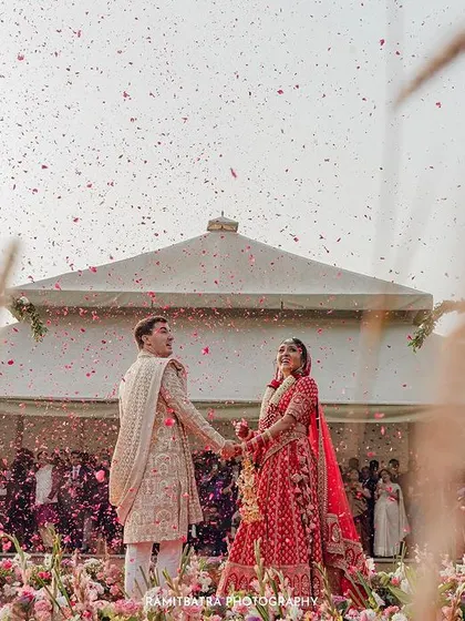 A wide shot of the Varmala ceremony, with the couple framed by pampas grass and the modern structure of the venue.