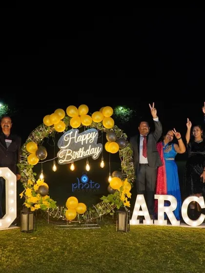 A celebratory group photo from an outdoor 60th birthday party at night, with everyone cheering in front of the large decorative signs.