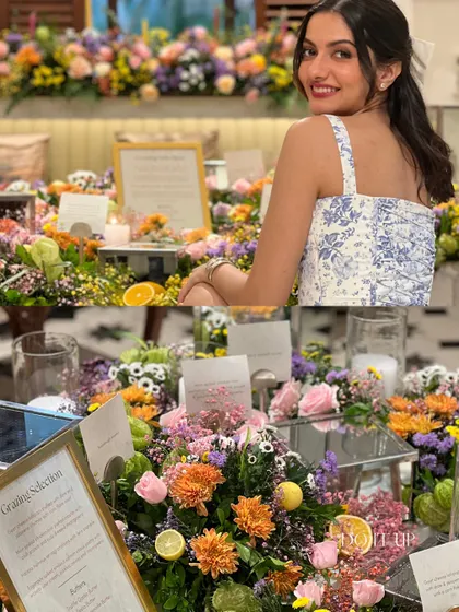 A vibrant grazing table overflowing with colorful flowers. This setup for an intimate gathering shows a woman smiling, surrounded by a beautiful arrangement of florals and personalized details.