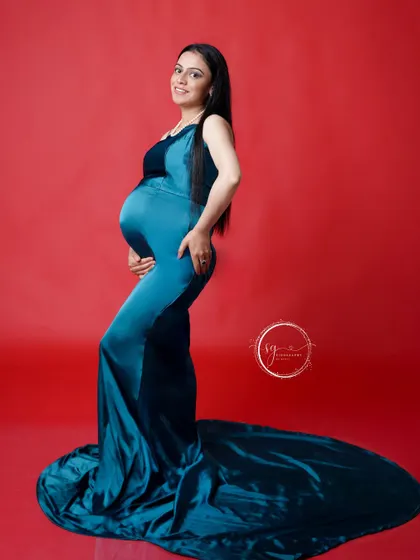 A confident side-profile portrait showcasing the baby bump. The sleek teal gown and bold red background create a modern and vibrant look.