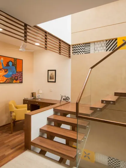 On the second floor of Kavyam Residence, an open study nook is tucked in neatly beside the staircase. It features a warm wooden desk, a cheerful yellow chair, and vibrant artwork, all illuminated by a skylight.