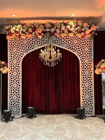 A royal photo booth or small stage setup, featuring a white jali arch against a rich maroon velvet curtain, topped with a floral arrangement and a chandelier.