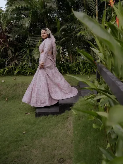 A bride in a flowing pink gown, captured from the side in a lush garden setting.