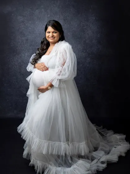 This full-length shot showcases the beautiful design of the white maternity gown. The soft ruffles and long train add a touch of glamour to this elegant studio portrait.