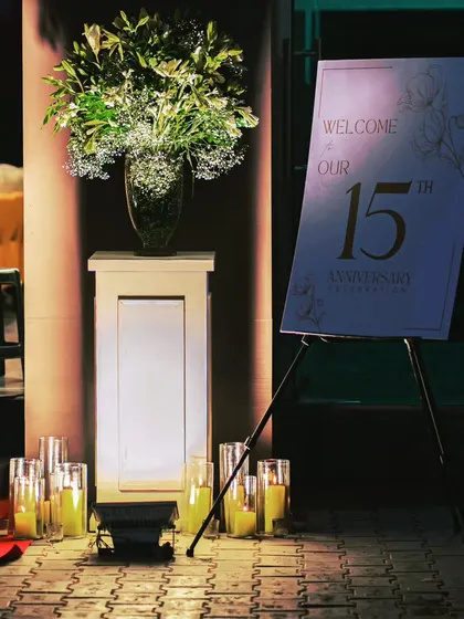 The welcome area for the 15th-anniversary event, featuring a custom sign, a large floral arrangement, and candle lighting to create an inviting entrance.
