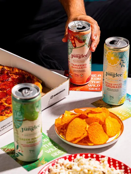 A lifestyle shot showing canned cocktails paired with pizza and snacks. This cross-promotional styling suggests perfect serving occasions to the consumer.