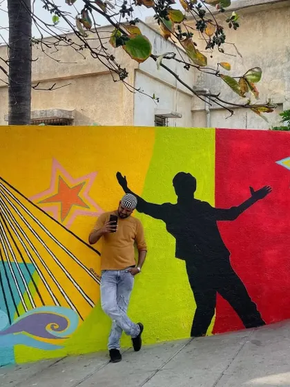 A fan poses against a colorful wall outside Shah Rukh Khan's house, "Mannat." The wall features a silhouette of SRK's iconic pose, making it a popular spot for fans to pay tribute to the Bollywood king.