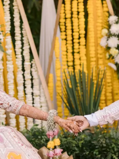 A close-up of the couple holding hands, showing the bride's beautiful henna and floral jewelry against the Haldi decor.