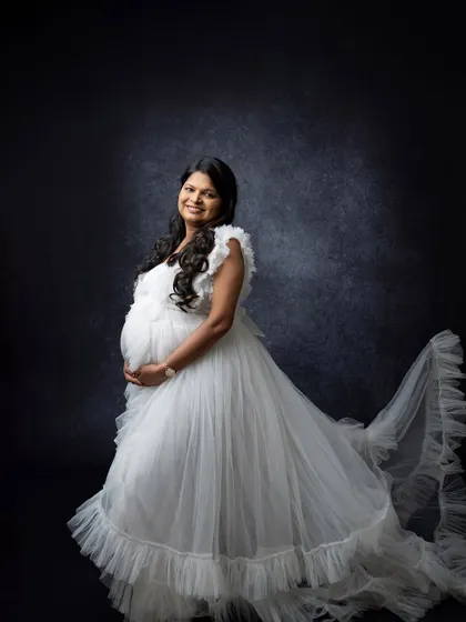 A solo maternity portrait in a flowing white gown against a dark studio background. The contrast creates a timeless and ethereal look, highlighting the mother's joyful expression.