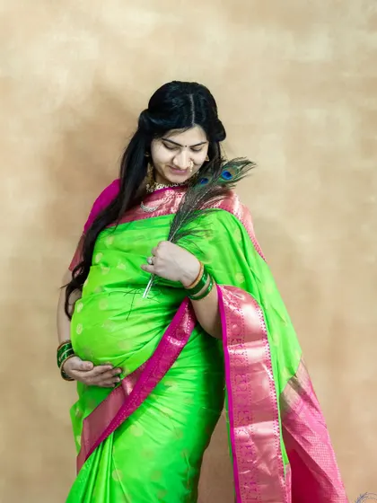 A traditional studio portrait of a mom-to-be in a green and pink silk saree, holding a peacock feather.