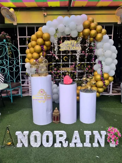 A birthday setup on a lawn with a white and gold balloon arch and marquee letters.