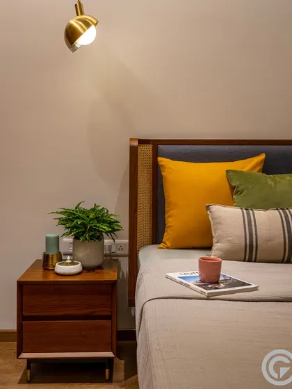 A close-up of a bedside table in a guest bedroom, showing the detail of the cane-paneled headboard and the mix of textures. Small details like a potted plant and a brass wall sconce complete the look.
