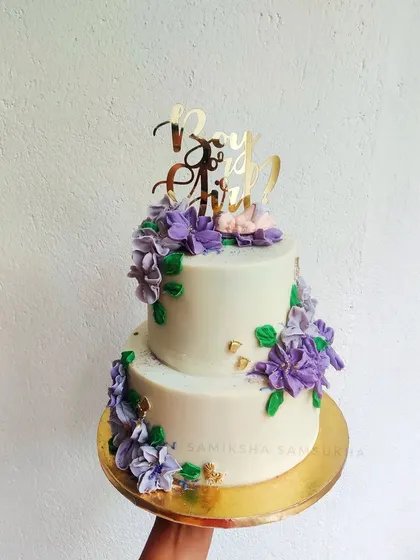 An elegant two-tiered baby shower cake finished entirely in ganache. The purple sugar flowers add a beautiful pop of color to the classic white design.