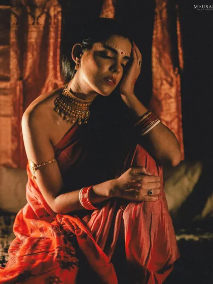 A full-body shot from the editorial series, showing the model in a seated, thoughtful pose. The warm, dramatic lighting highlights the texture of the saree and her graceful form.