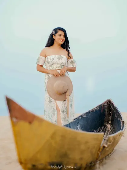 A beautiful solo portrait of the bride-to-be standing on the beach next to a rustic boat, holding a sun hat.