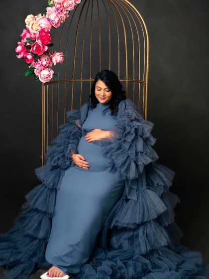 A regal portrait of a mom-to-be in our blue ruffled gown, seated in a golden birdcage prop for a whimsical and enchanting look.