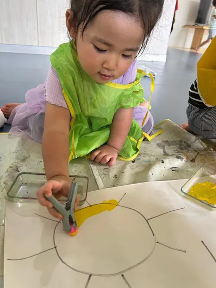 A toddler in a green apron is completely absorbed in painting a sun. We encourage this deep focus, which is a key part of play-based learning.