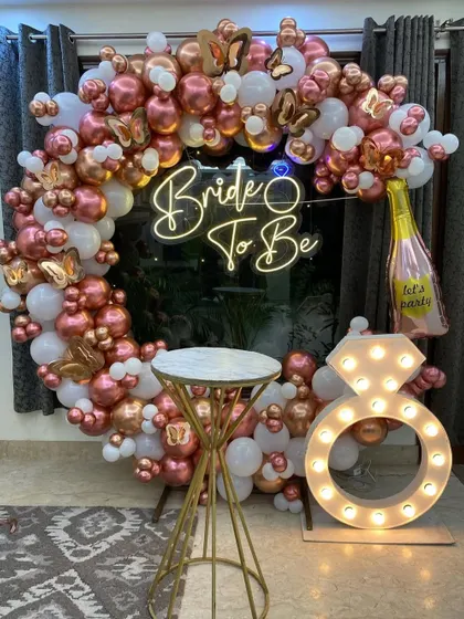 I design trendy and fun photo booths for pre-wedding events. This 'Bride to Be' setup includes a custom neon sign, a circular balloon arch in rose gold and white, and large marquee props like a light-up diamond ring.