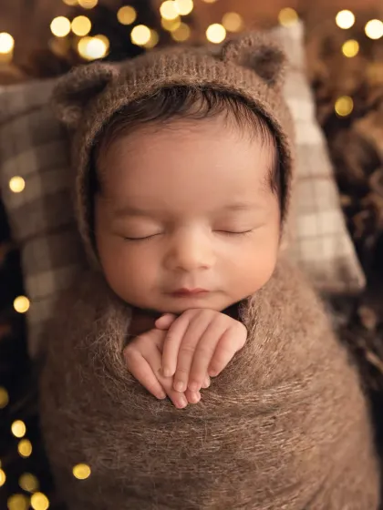 A close-up of a newborn boy in a brown swaddle and bear bonnet, with soft, warm lights twinkling in the background.