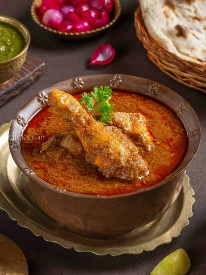 A classic Chicken Curry with a leg piece, served in a traditional brass bowl with pickled onions and naan.