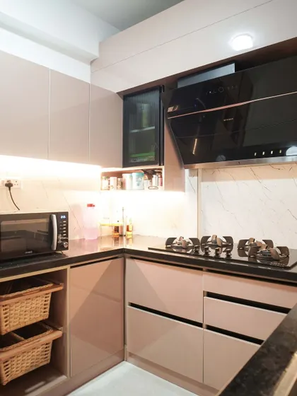A U-shaped kitchen designed for a 3BHK apartment, featuring beige handleless cabinets and a black countertop. The layout includes open wicker basket storage and a dedicated space for the microwave.