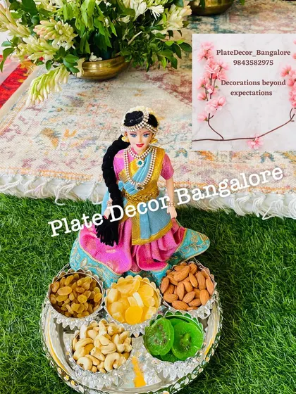 A modern take on the seer platter, this arrangement features a Barbie doll dressed in a traditional saree, holding a tray of dry fruits.
