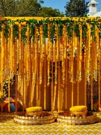 A classic Haldi seating arrangement with a backdrop of marigold garlands against yellow drapes. The two traditional 'peedas' (low stools) are ready for the bride and groom.