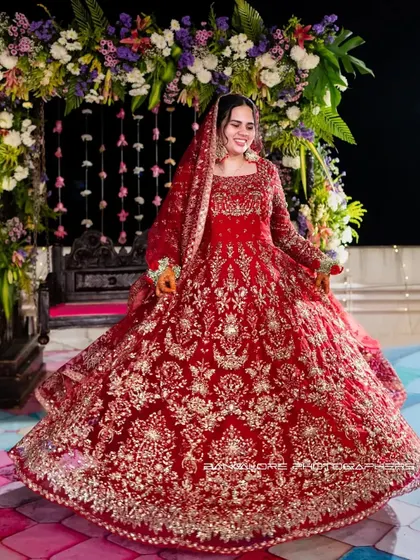 The bride, seated and smiling, in her magnificent red reception outfit. The floral backdrop and her joyful expression create a picture of pure celebration.