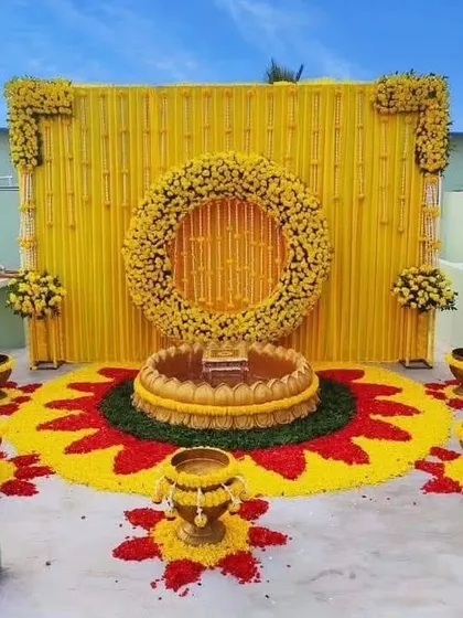 We can create beautiful setups even on a terrace. This Haldi theme features a backdrop of yellow drapes and a floral ring, with a colorful rangoli made of flower petals on the floor.