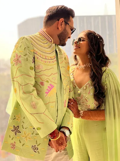 A loving glance between a couple dressed in our coordinated carnival-themed outfits. The intricate, colorful embroidery on his sherwani beautifully matches the floral details on her lime green jumpsuit.
