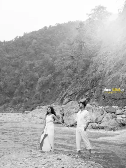 A timeless black and white photograph of a couple standing by the river, with the sun creating a beautiful flare over the water and mountains.