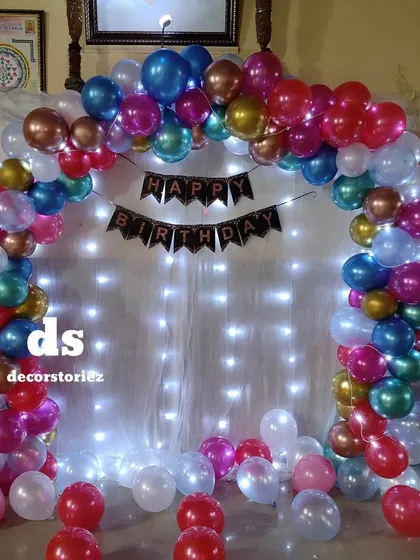 A vibrant and festive balloon arch using multi-colored chrome balloons against a backdrop of white curtains and fairy lights, perfect for any lively celebration.