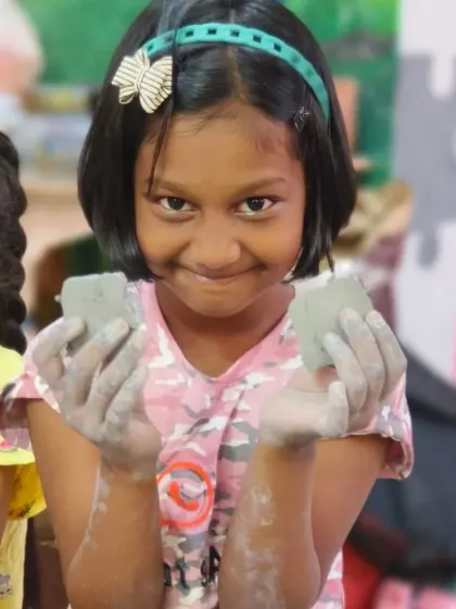 A girl grins as she holds up two pieces of clay, ready to start her next project. The enthusiasm of our young students is infectious.