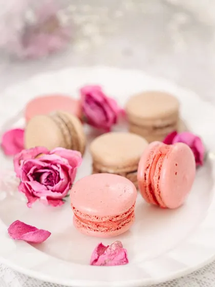 A close-up of a pink macaron, with other macarons and flowers softly blurred in the background.